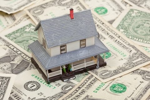 A model house sits on top of several scattered one-dollar bills, representing the concept of home finances or real estate investment.