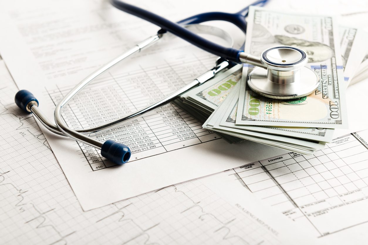 A stethoscope rests on a stack of US dollar bills atop medical charts and documents.