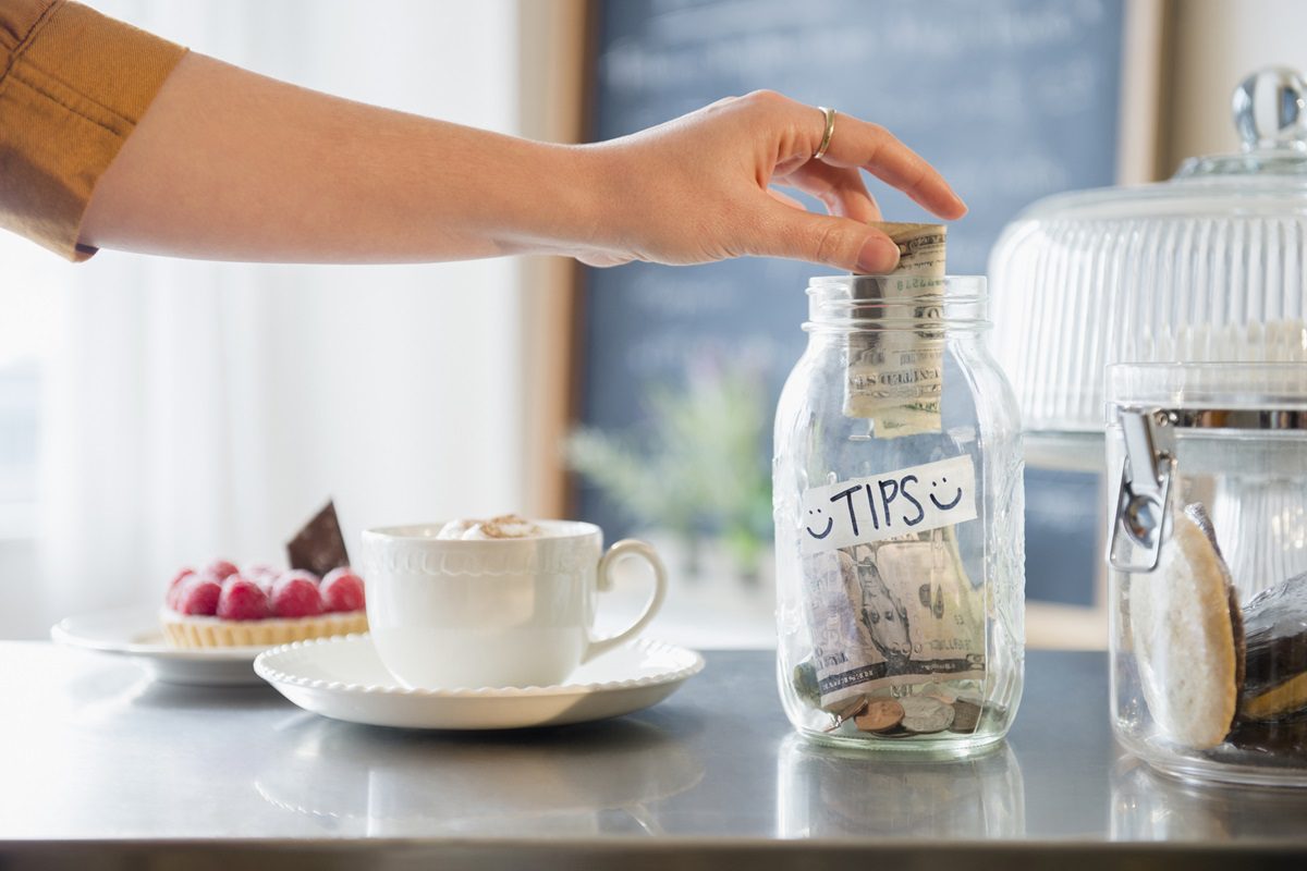 A person places cash into a glass tip jar labeled TIPS on a counter next to a cup of coffee, a dessert, and another jar containing cookies.