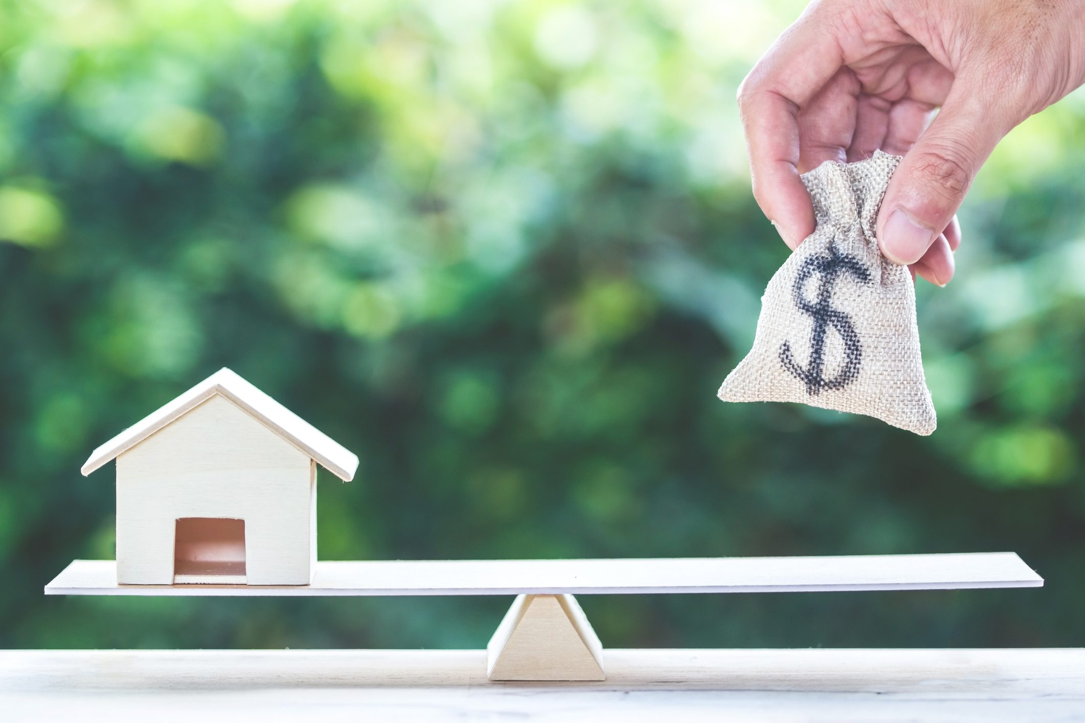 A small wooden house and a hand holding a money bag are balanced on opposite ends of a seesaw, symbolizing housing costs or mortgage.
