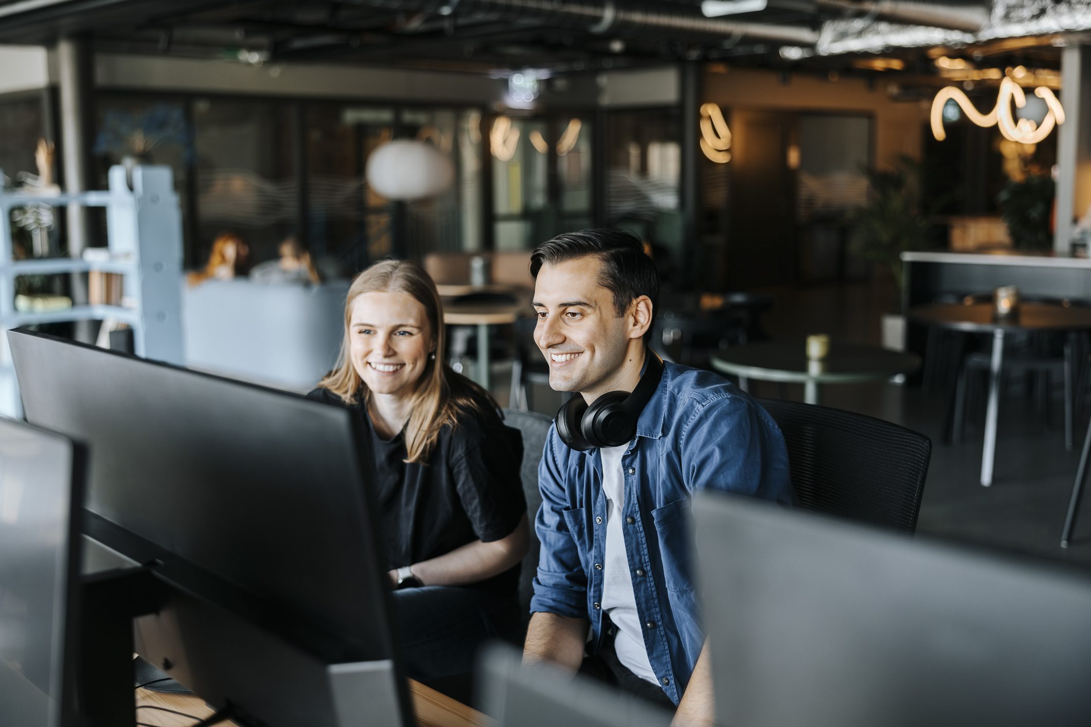 Two colleagues sit side by side at desks, looking at computer monitors and smiling in a modern open-plan office space.