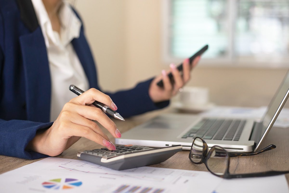 A person in business attire uses a calculator, holds a smartphone, and works at a laptop with financial charts and eyeglasses on the desk.
